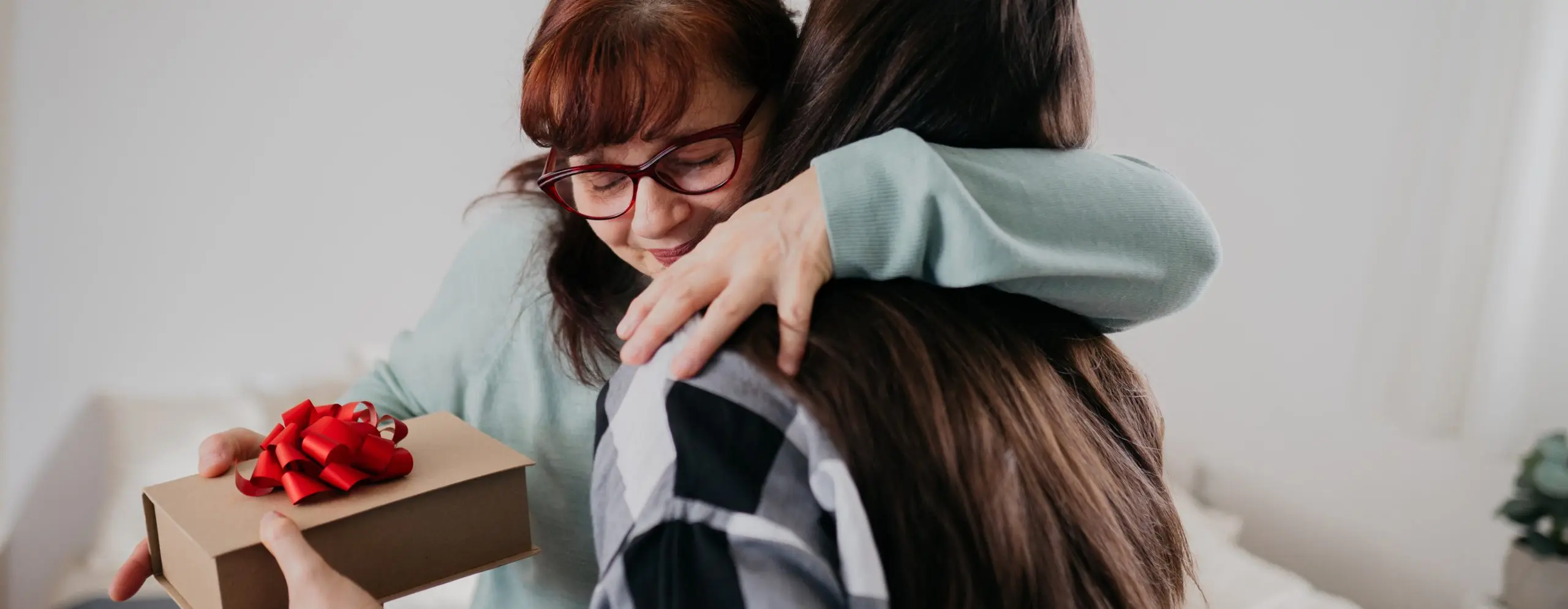 Mulher entrega um presente embrulhado para sua mãe, que a abraça emocionada em um gesto de carinho pelo Dia das Mães.