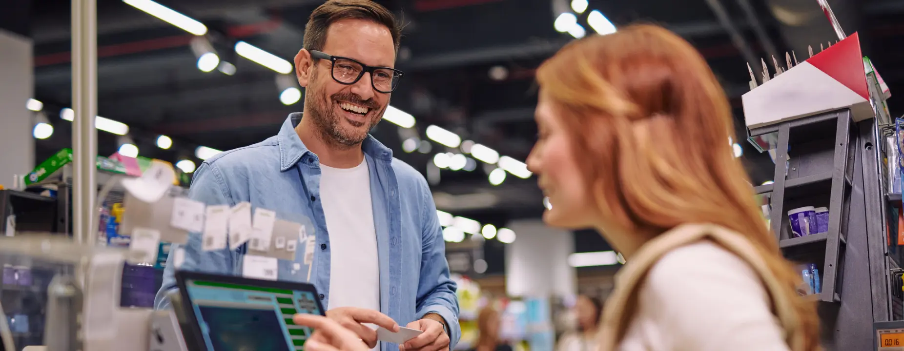 Homem sorrindo no caixa do supermercado, entregando um cartão de pagamento ou voucher para a atendente.