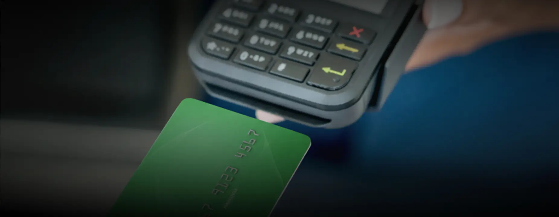 Uma mão segurando um cartão verde com números parcialmente visíveis, aproximando-o de uma máquina de cartão preta para realizar um pagamento por aproximação.