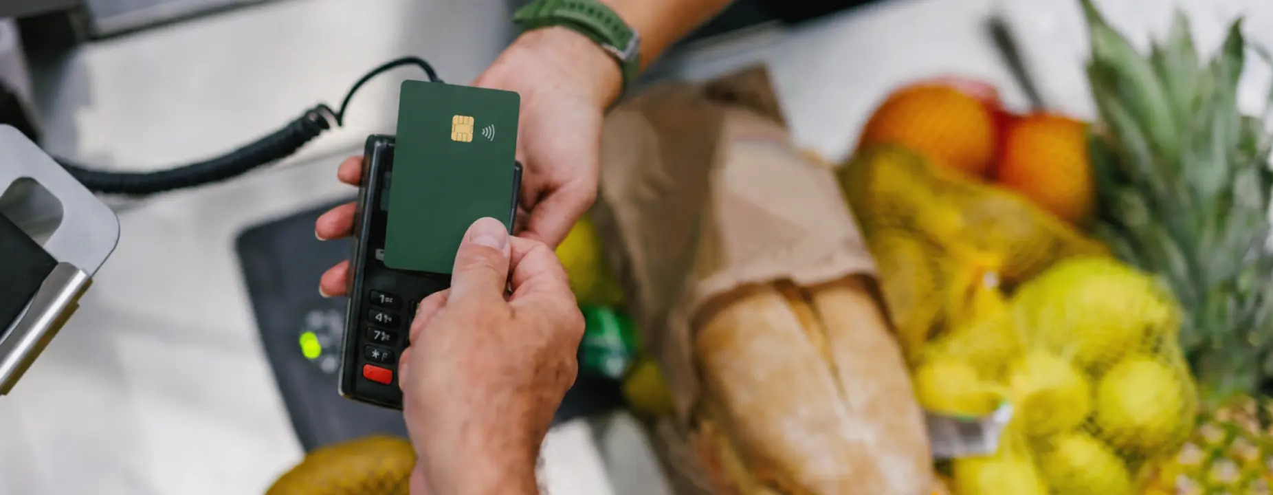 Pessoa realizando pagamento por aproximação com cartão de crédito verde escuro em uma maquininha de cartão no caixa de supermercado, com frutas, pão e compras ao fundo.